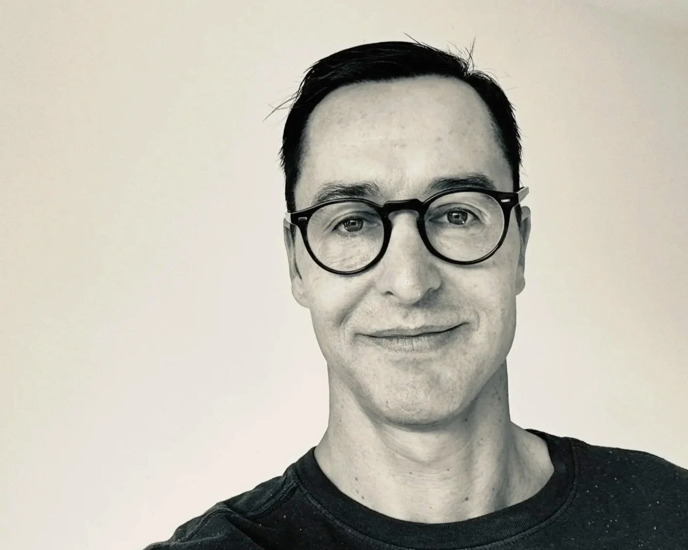 Black-and-white portrait of a man wearing round glasses and a dark shirt, smiling slightly, with a plain light-colored background.