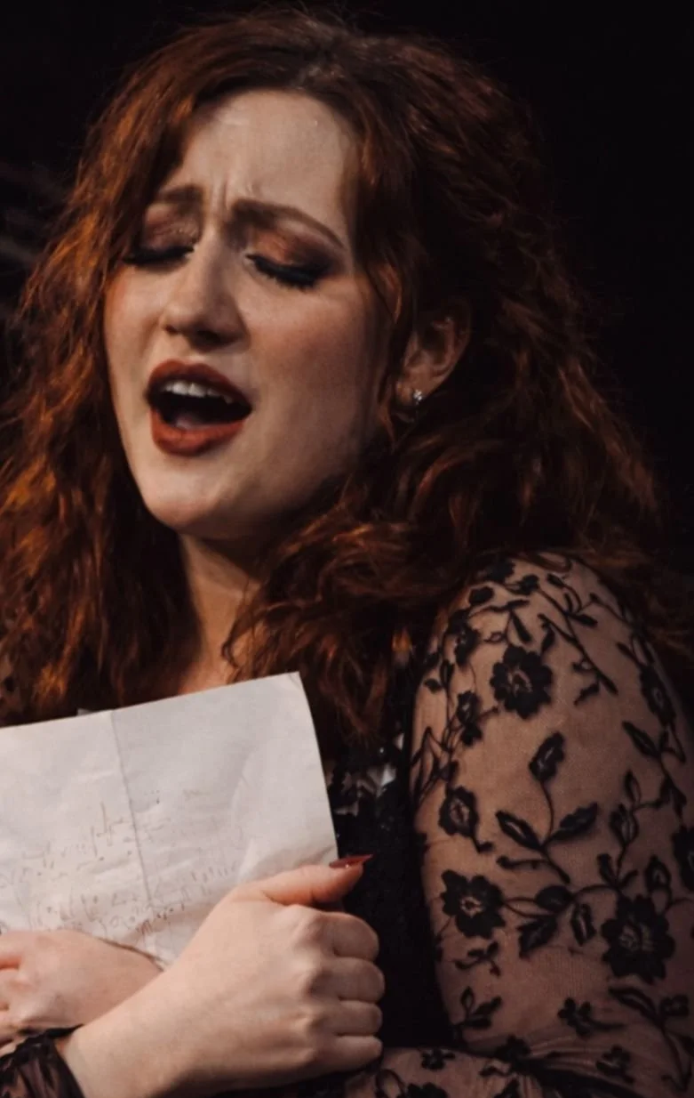 A woman with red curly hair singing passionately, holding a piece of paper, wearing a black lace top with floral patterns.