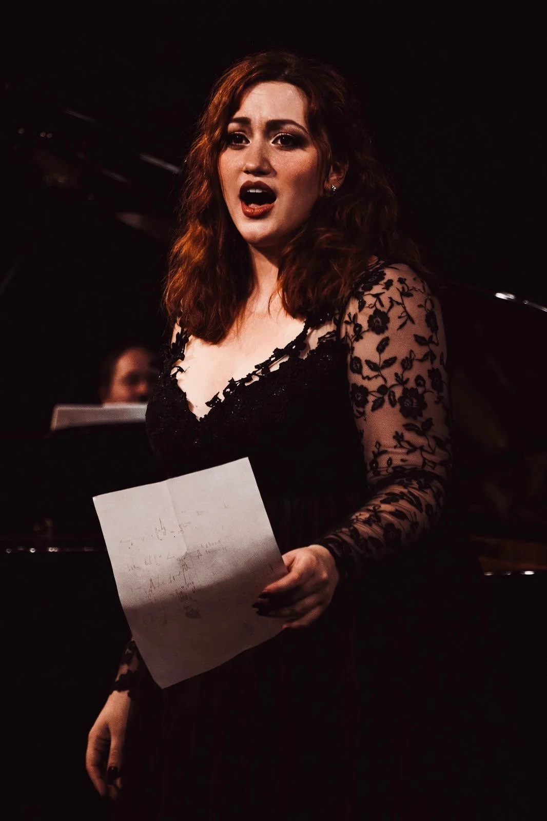 A woman with red hair wearing a black lace dress, singing or speaking with an expressive face, holding a sheet of paper, with a pianist in the background.