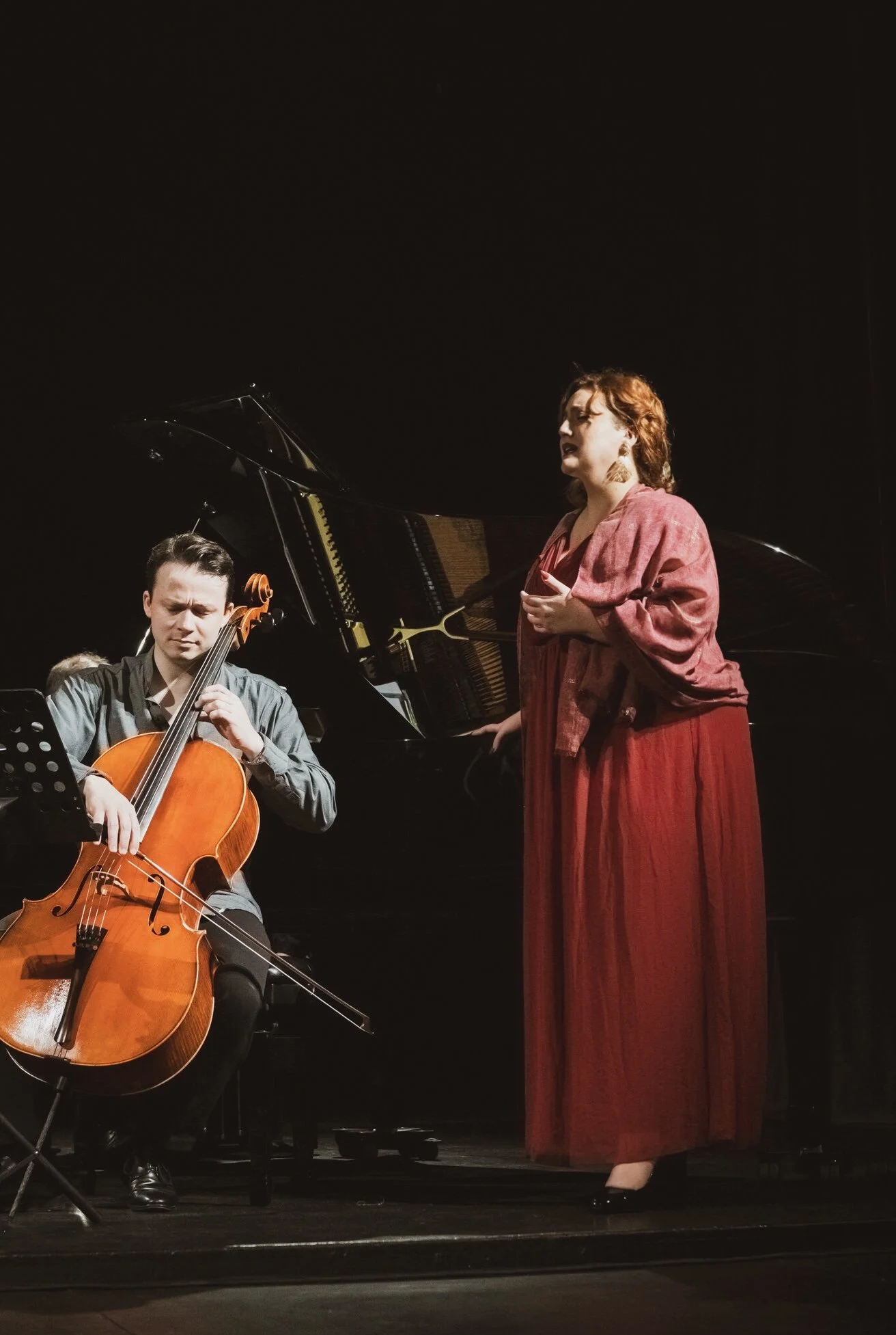 A woman singing with a closed eyes expression, standing next to a pianist and a cellist during a performance on a stage with a black background.