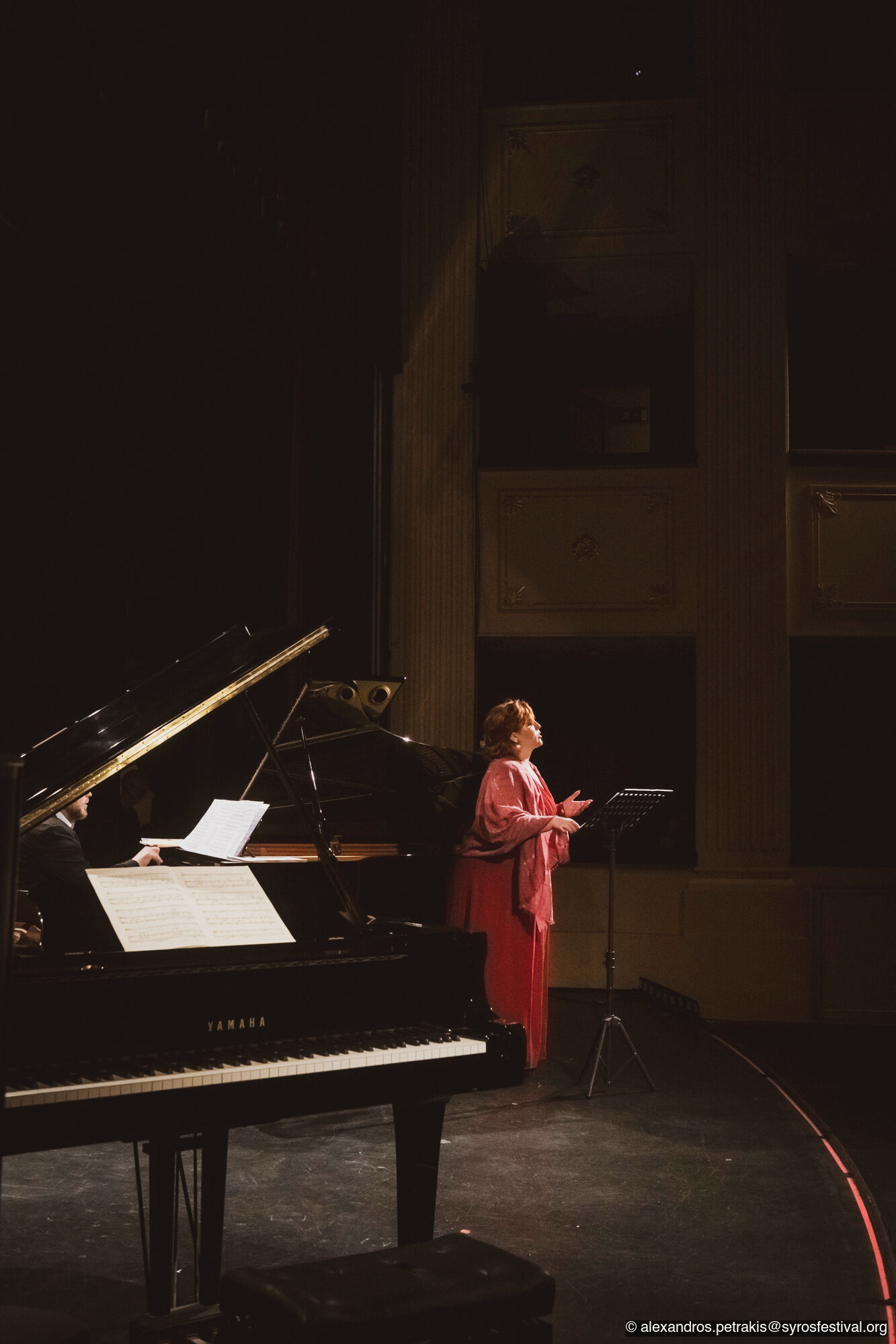 A woman in a red dress singing on a stage with a piano and sheet music, in a dimly lit theater or concert hall.