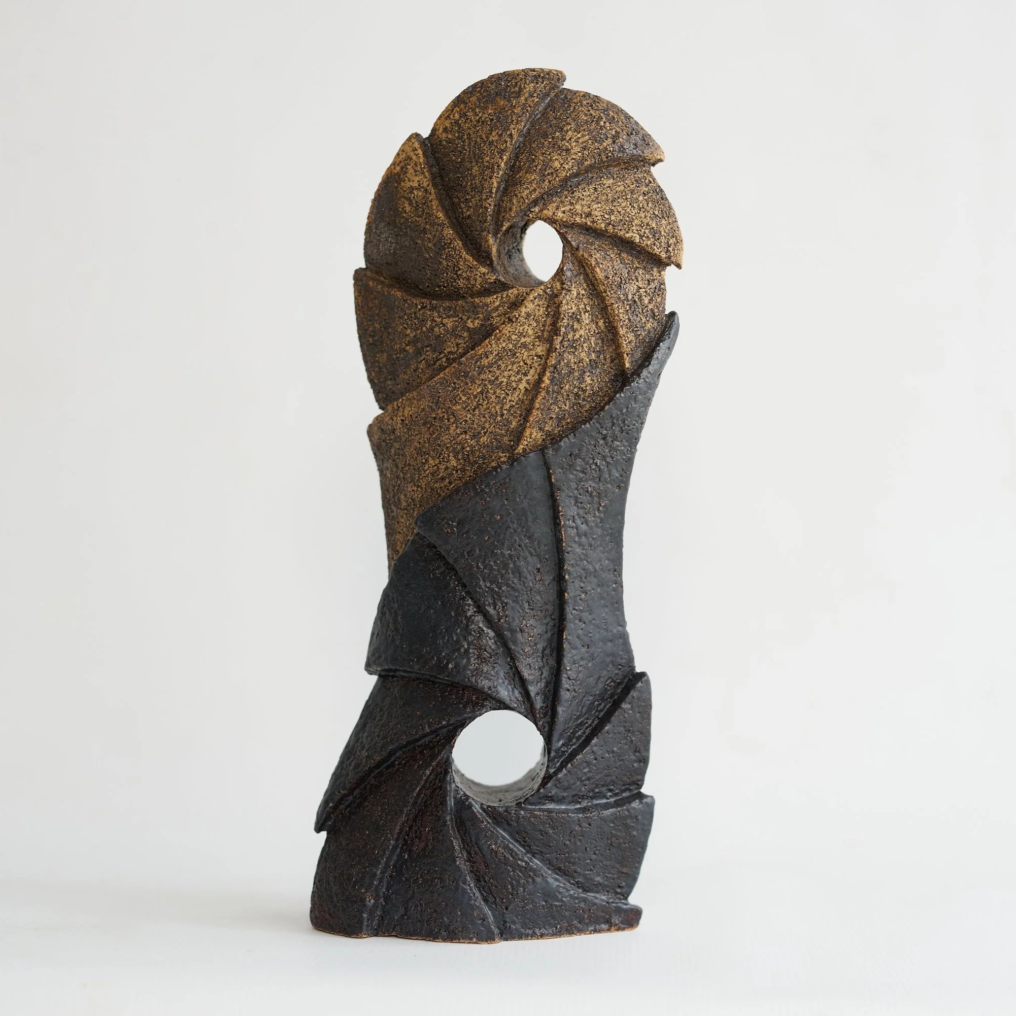 A carved ceramic sculpture with a textured, dark surface featuring a curling, spiral design against a plain white background.