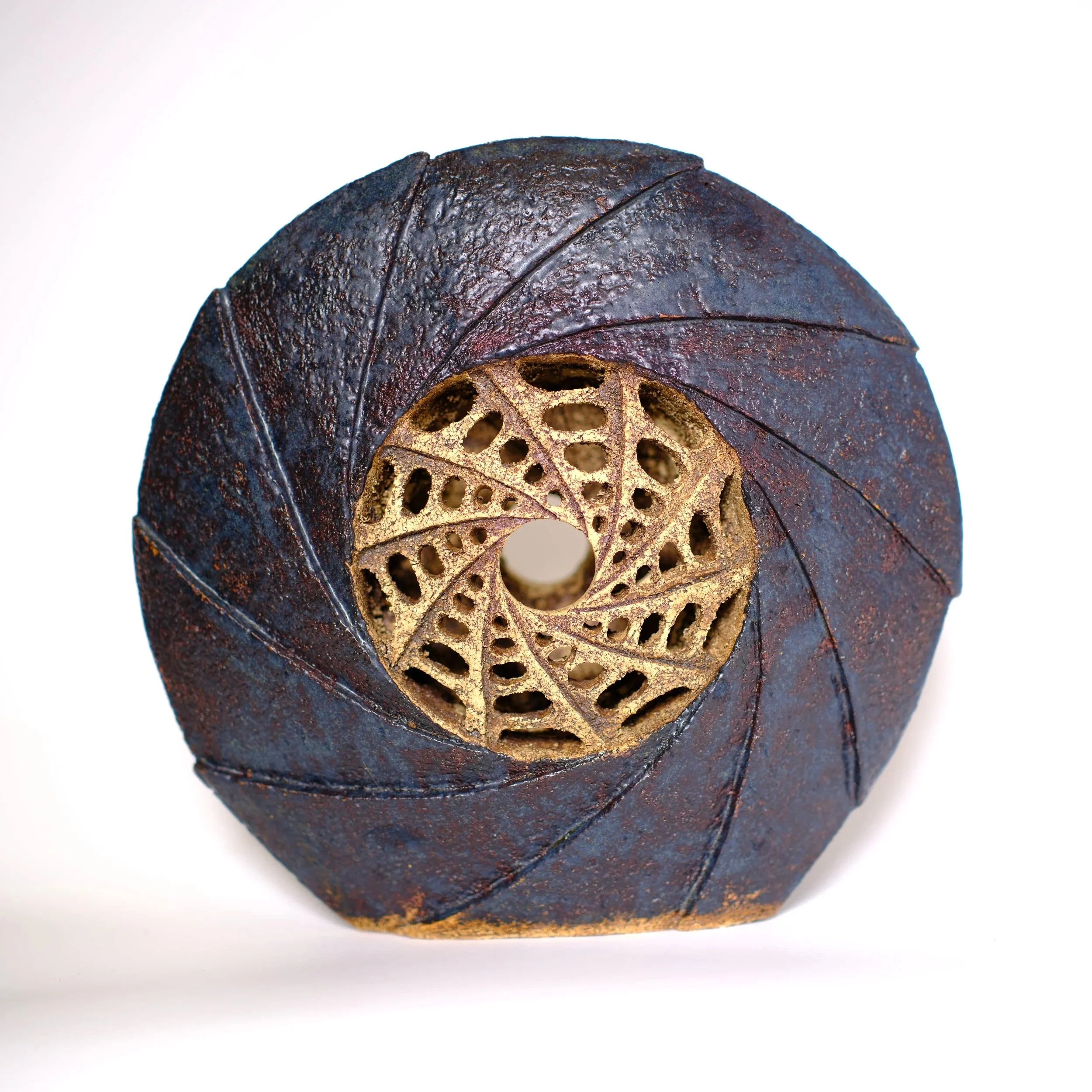 A round, carved ceramic sculpture featuring an outer ring of dark blue, with a clockwise spiralling design, and a pale beige inner circle featuring an anticlockwise spiralling design, with small oval holes carved through the hollow sculpture.