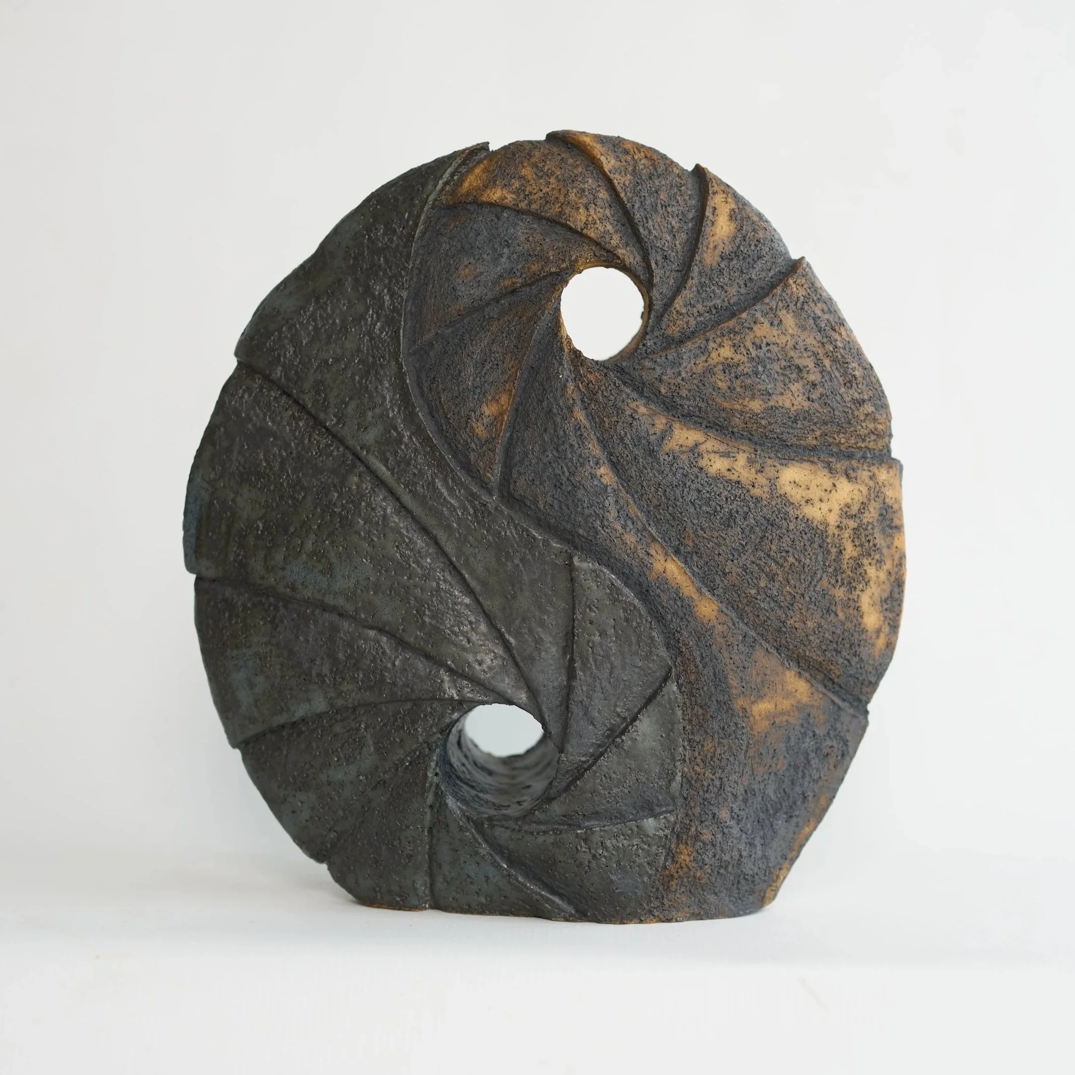 A round ceramic sculpture with two holes, featuring textured, dark surfaces with lighter patches, set against a plain white background.