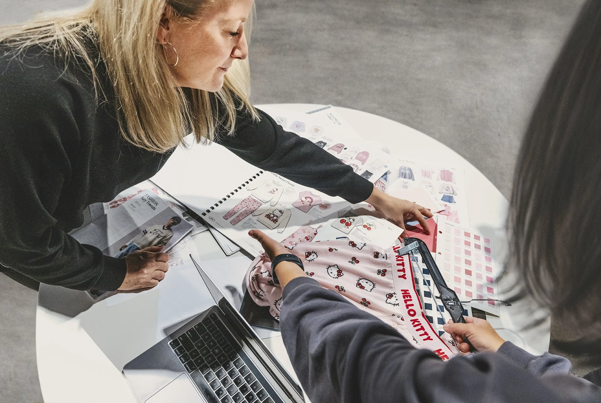 Groupe de personnes discutant de tissus avec des motifs Hello Kitty, une d'entre elles tient un échantillon de tissu, autour d'une table avec un ordinateur portable et des catalogues.