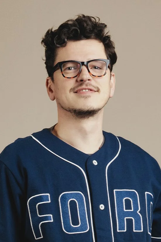 A young man wearing glasses and a blue sweater with the letters 'F O R' on it, smiling slightly.