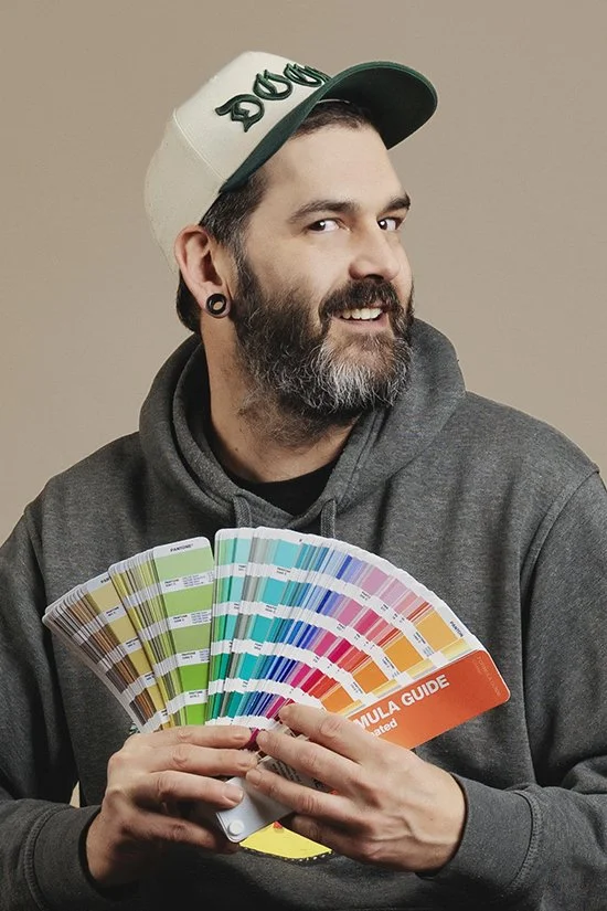 A smiling man with a beard wearing a white beanie with the word "DOG" on it and a gray jacket, holding a stack of books in standard colors.