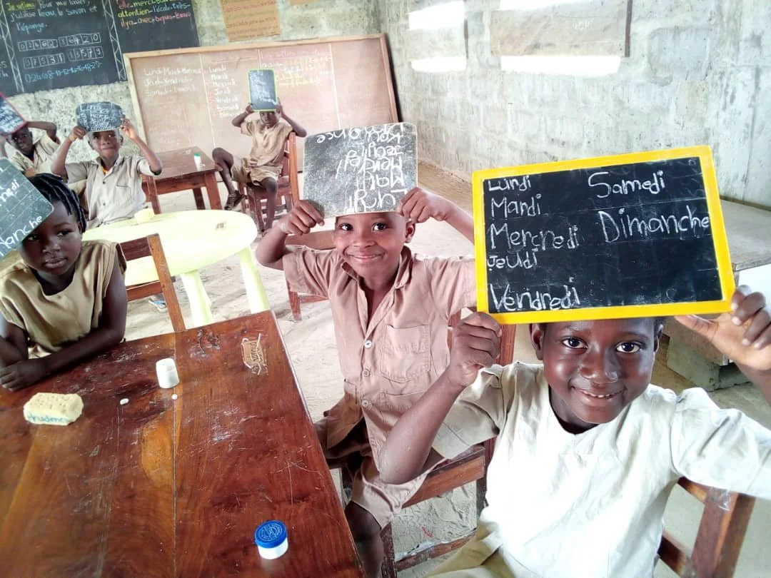 Groupe d'enfants en classe, certains portant des ardoises sur la tête avec des inscriptions, salle de classe simple avec tableau noir et chaises en bois.
