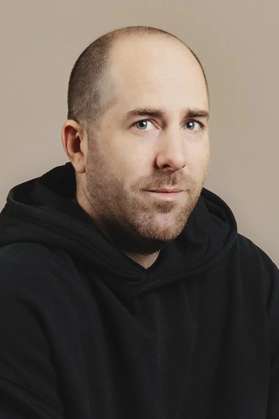 A man with a beard, wearing a black hoodie, looking at the camera, against a beige background.