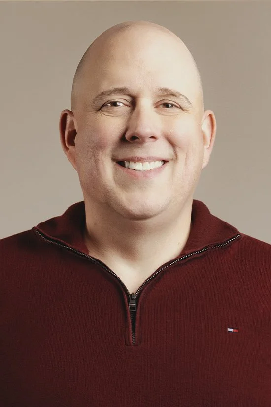 A smiling man with fair skin, wearing a dark brown zip-up sweater with a beige lining.