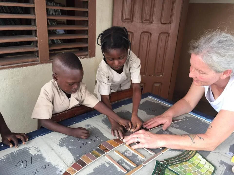 Une femme, probablement une éducatrice ou une enseignante, travaille avec deux enfants africains dans une salle de classe ou un centre éducatif. Elle leur montre des échantillons de carreaux de céramique ou de mosaïque sur une table.