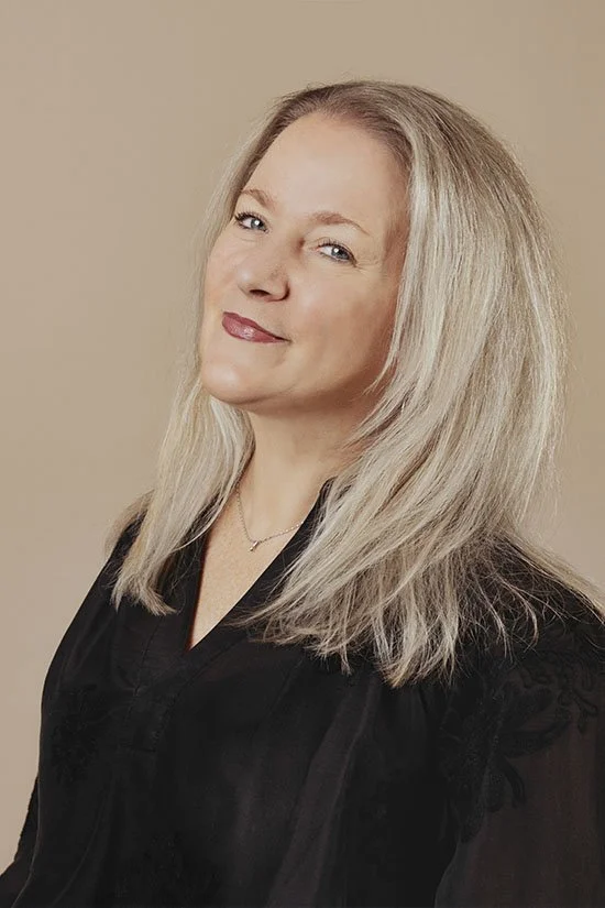 A woman with long blonde hair, wearing a black dress and smiling slightly, against a beige background.