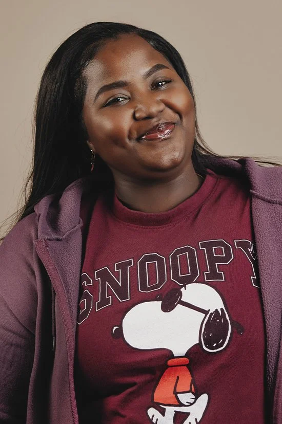 A smiling woman is wearing a burgundy Snoopy T-shirt and a purple jacket against a neutral background.