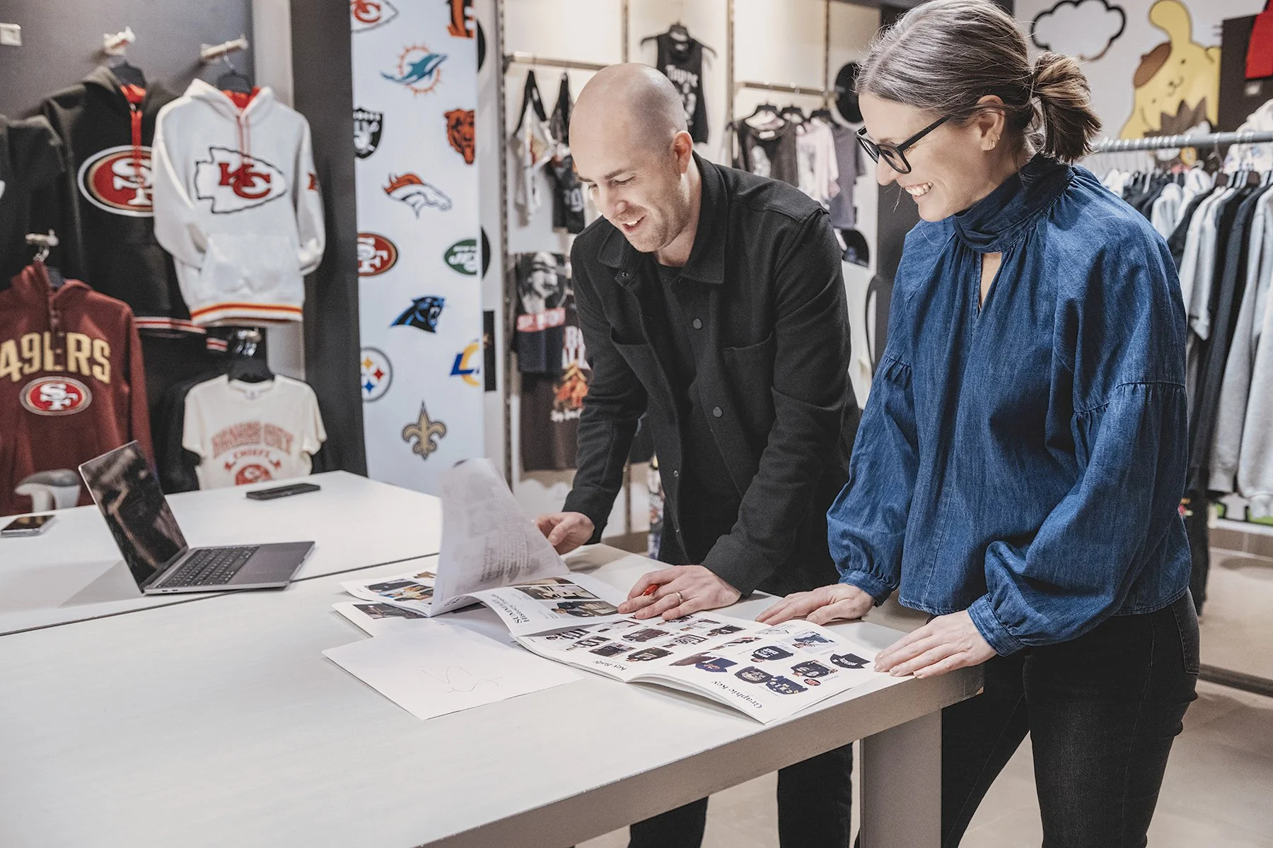 Deux personnes regardent des magazines ou catalogues sur une table dans une boutique de vêtements, avec des vêtements de sport et des logos NFL en arrière-plan.