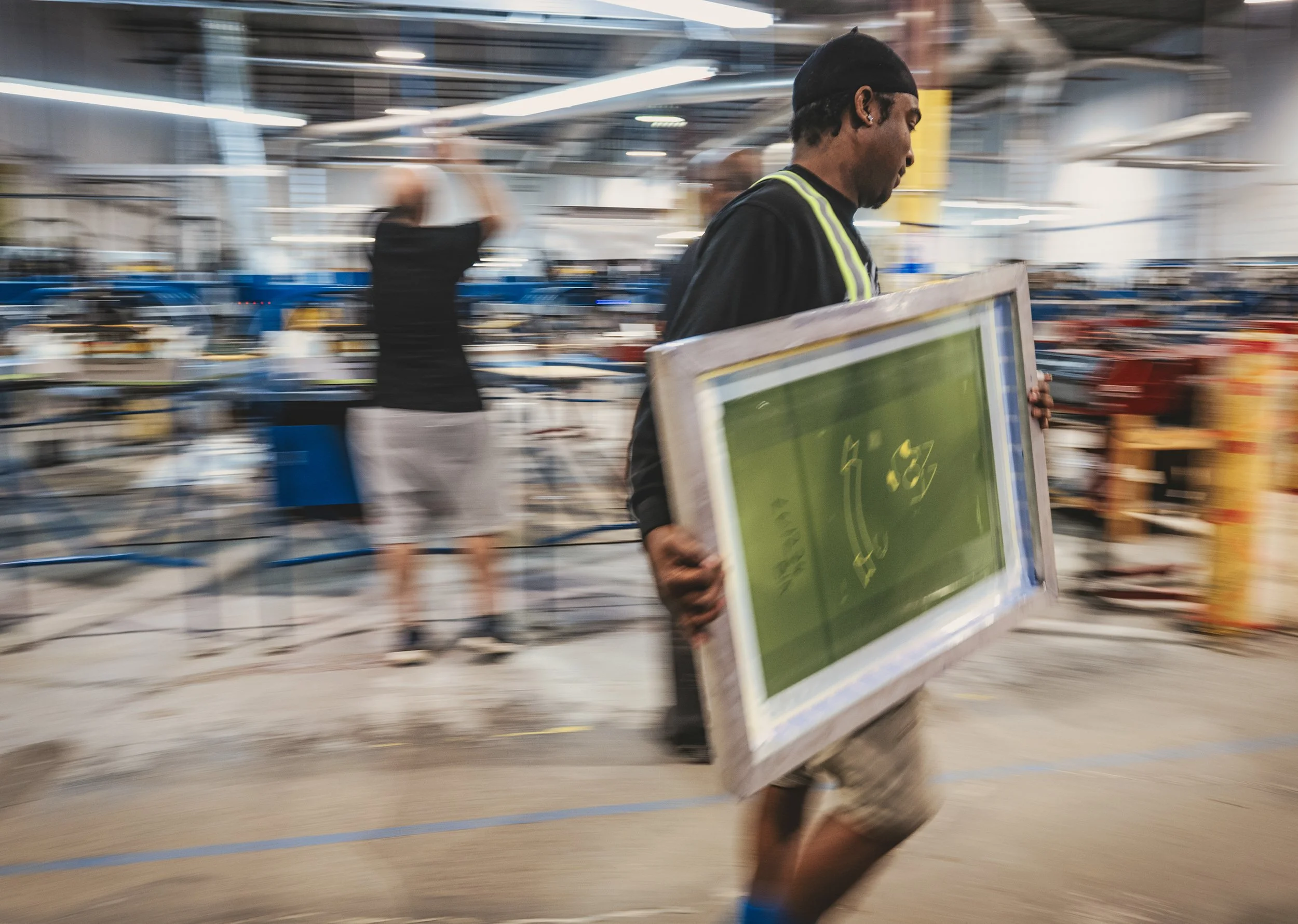 Une personne marche dans un centre commercial ou une grande surface, portant un tableau ou un panneau vert et noir. Le mouvement est vif, avec un flou artistique et un arrière-plan éclairé et animé.