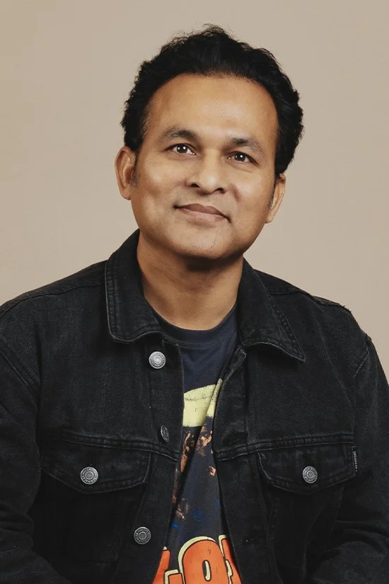 Photo of a smiling man wearing a black denim jacket and a graphic T-shirt, posing in front of a plain background.