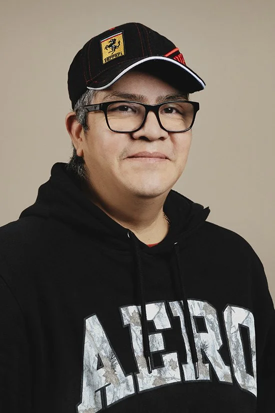 A man wearing a Ferrari cap, sunglasses, and a black hoodie, standing in front of a beige wall.