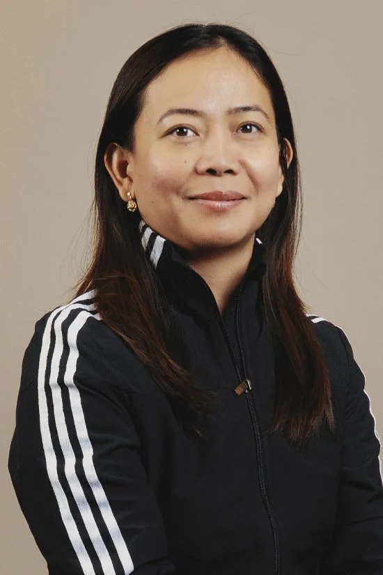 A smiling woman wearing a black sports jacket with white stripes, with long brown hair and gold earrings, against a neutral background.