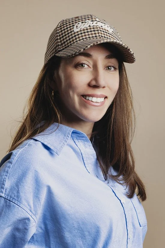 A smiling woman wearing a checkered cap and a light blue shirt, against a beige background.