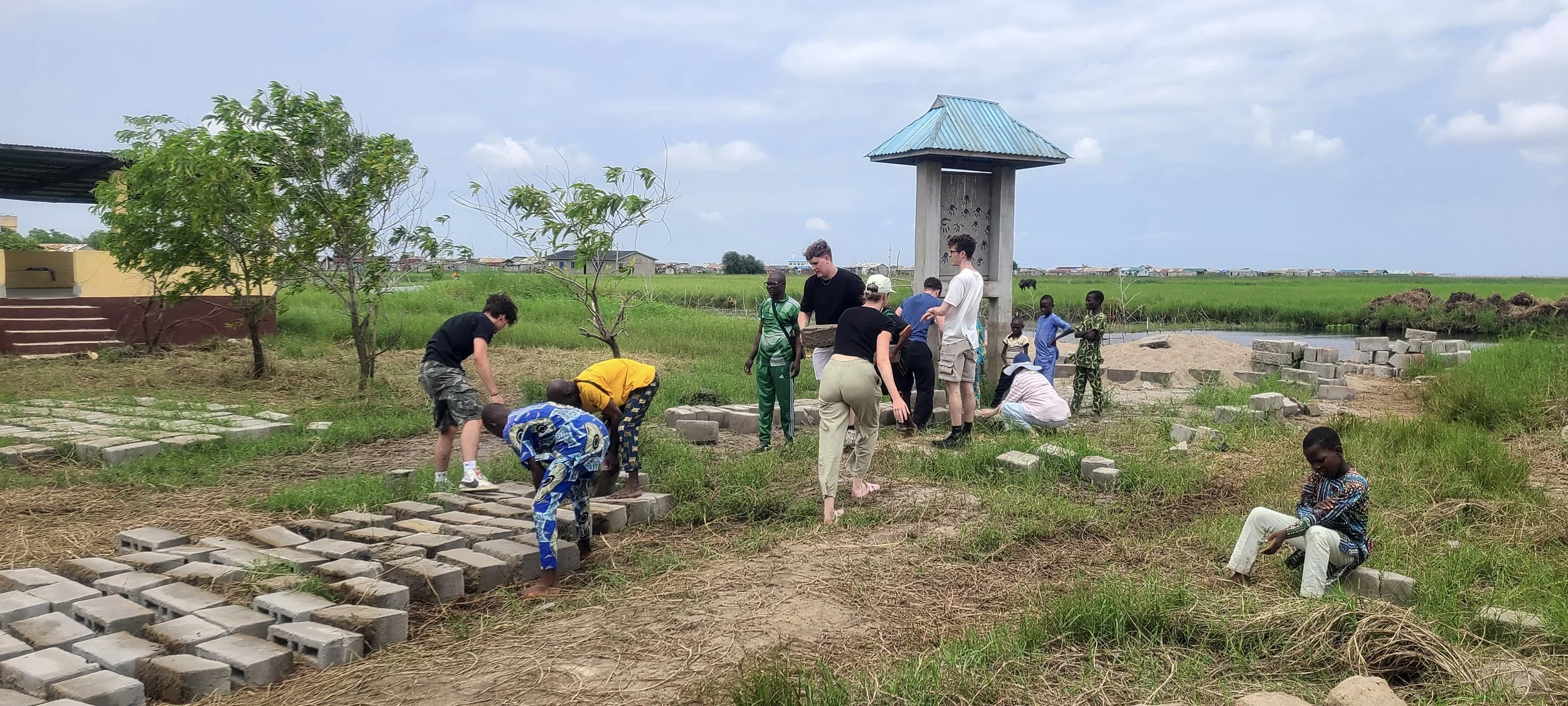 Groupe de personnes construisant une structure en briques dans un espace extérieur, avec un paysage rural en fond.