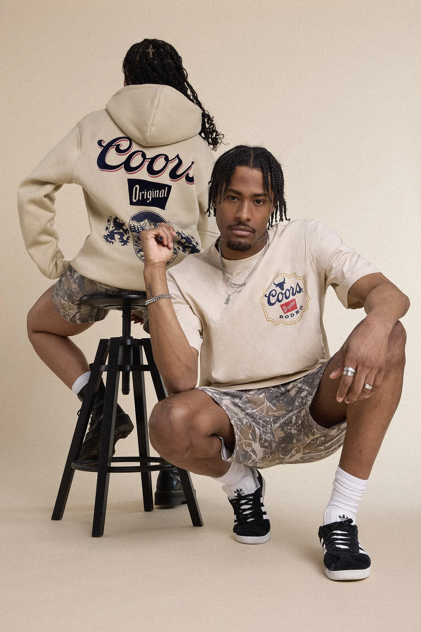 Deux jeunes hommes posent dans un studio avec un arrière-plan beige. L'un est assis sur un tabouret, portant un t-shirt beige avec un logo Coors, des shorts camouflage, un bracelet, une chaîne et une bague, avec des baskets noires et des chaussettes blanches. L'autre est debout, comme posé sur le tabouret, portant une sweat-shirt beige avec un logo Coors, des shorts camouflage, et des baskets noires. L'homme assis tient une tournevis dans sa main, tandis que celui derrière regarde fixement l'objectif.