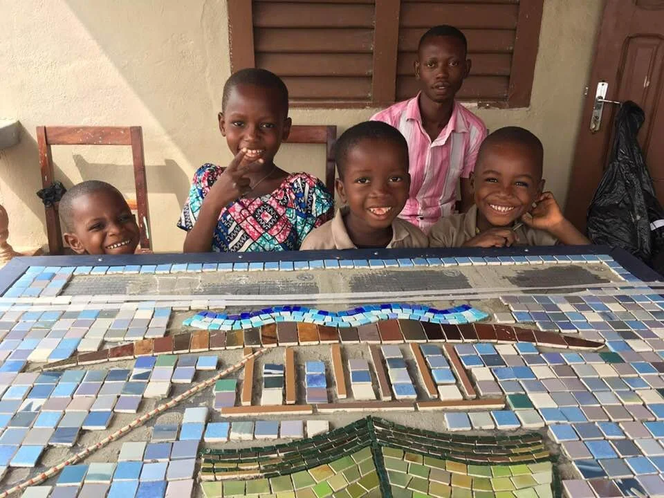 Groupe de cinq enfants africains souriants posant derrière une table de mosaïque colorée, dans une pièce avec une fenêtre en bois et un mur beige.
