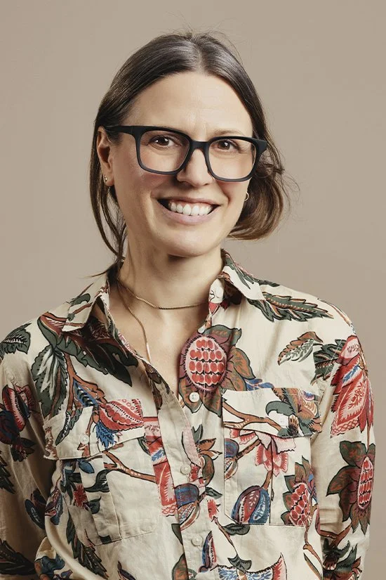 Portrait of a woman wearing black glasses and a colorful floral-print shirt, smiling against a beige background.