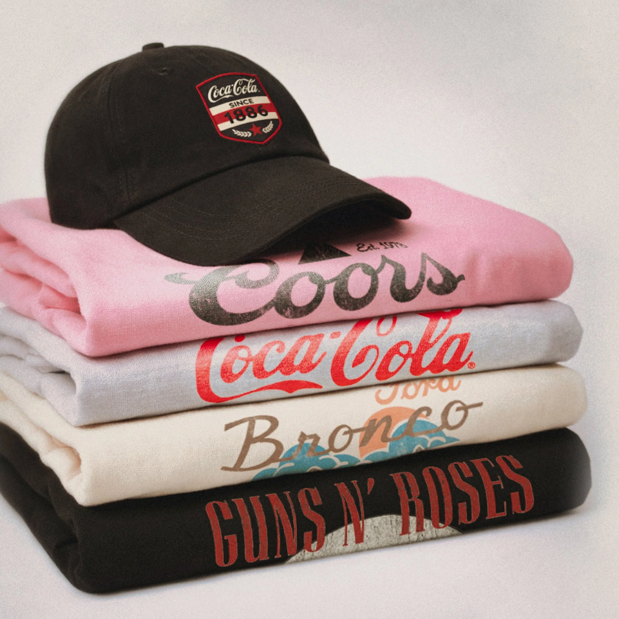 Une casquette noire avec un patch rouge, blanc et noir de Coca-Cola 1886, posée sur un paquet de vêtements de couleur rose.