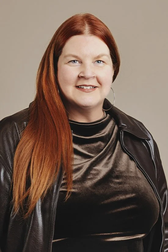A woman with long red hair is wearing a black leather jacket and a gray satin top; she is smiling against a neutral background.