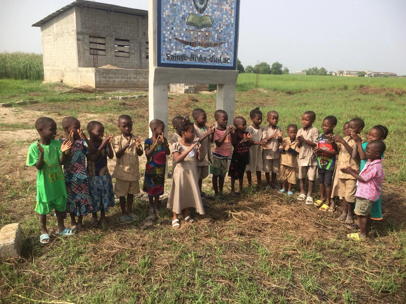 Groupe d'enfants africains en cercle, certains avec des vêtements colorés, dans un champ de terre avec un bâtiment en construction derrière eux.