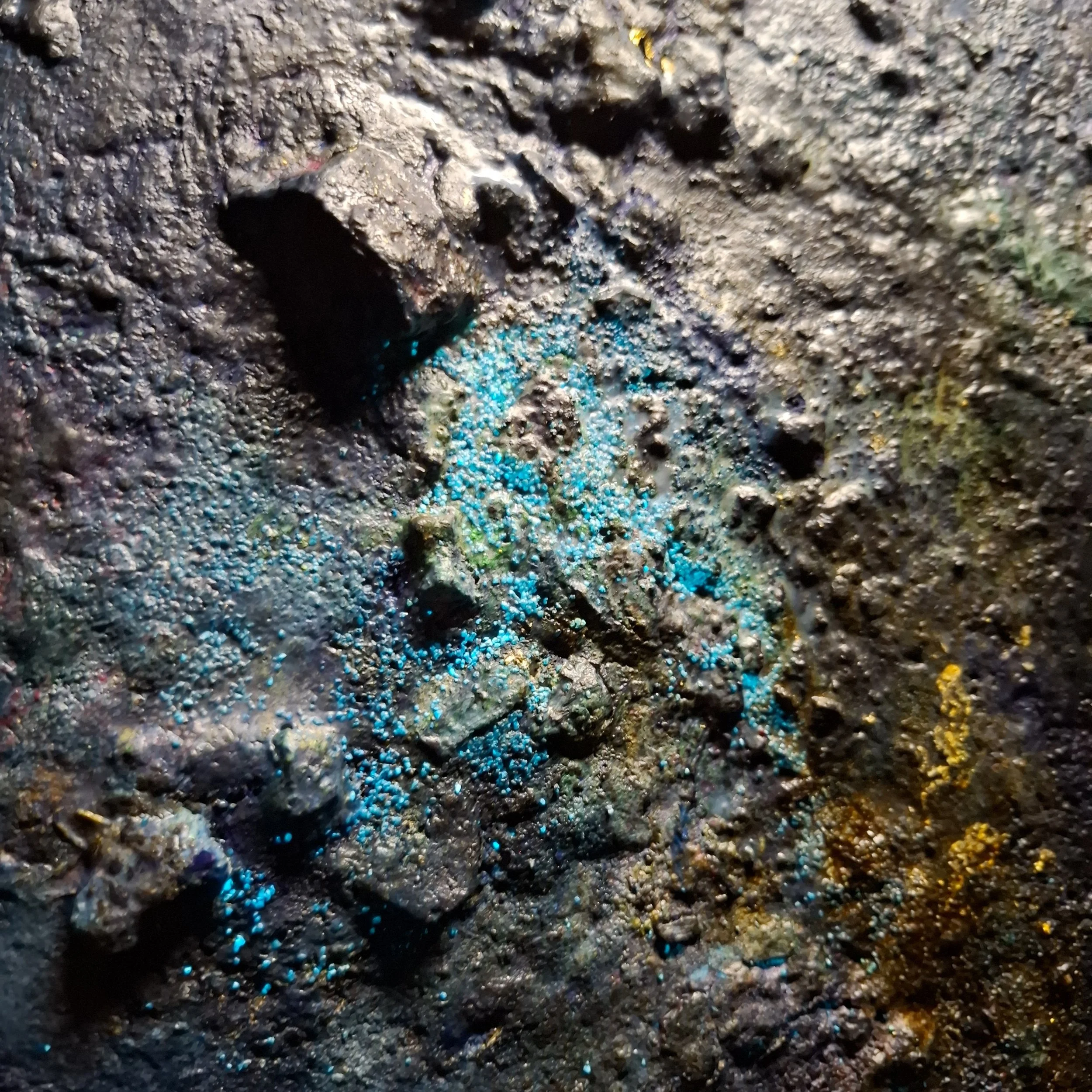 Naheaufnahme von einer rauen, schwarzen Oberfläche mit kleinen, schillernden blauen und bunten Kristallen oder Partikeln
