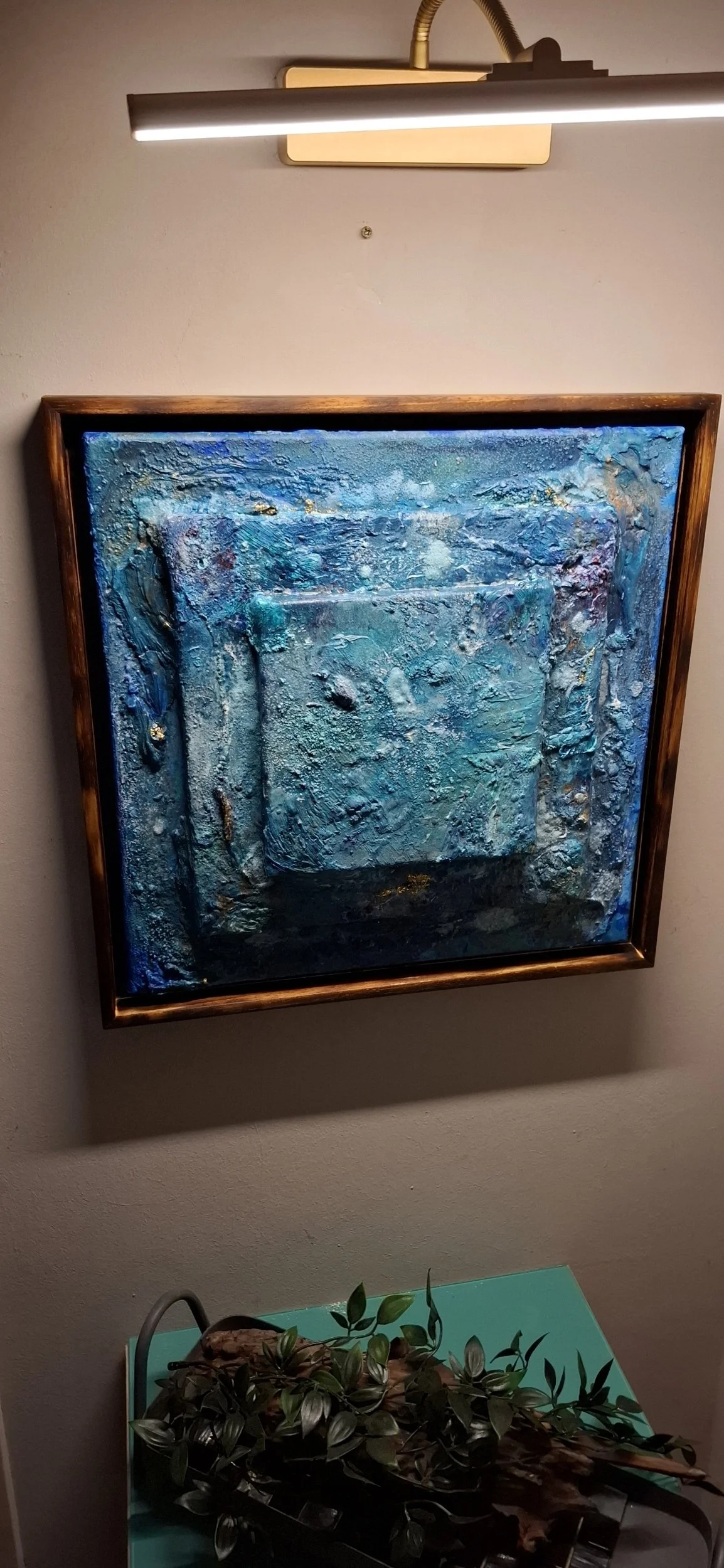 Abstraktes, blaues Gemälde mit dicker Textur in einem Holzrahmen, beleuchtet von oben.