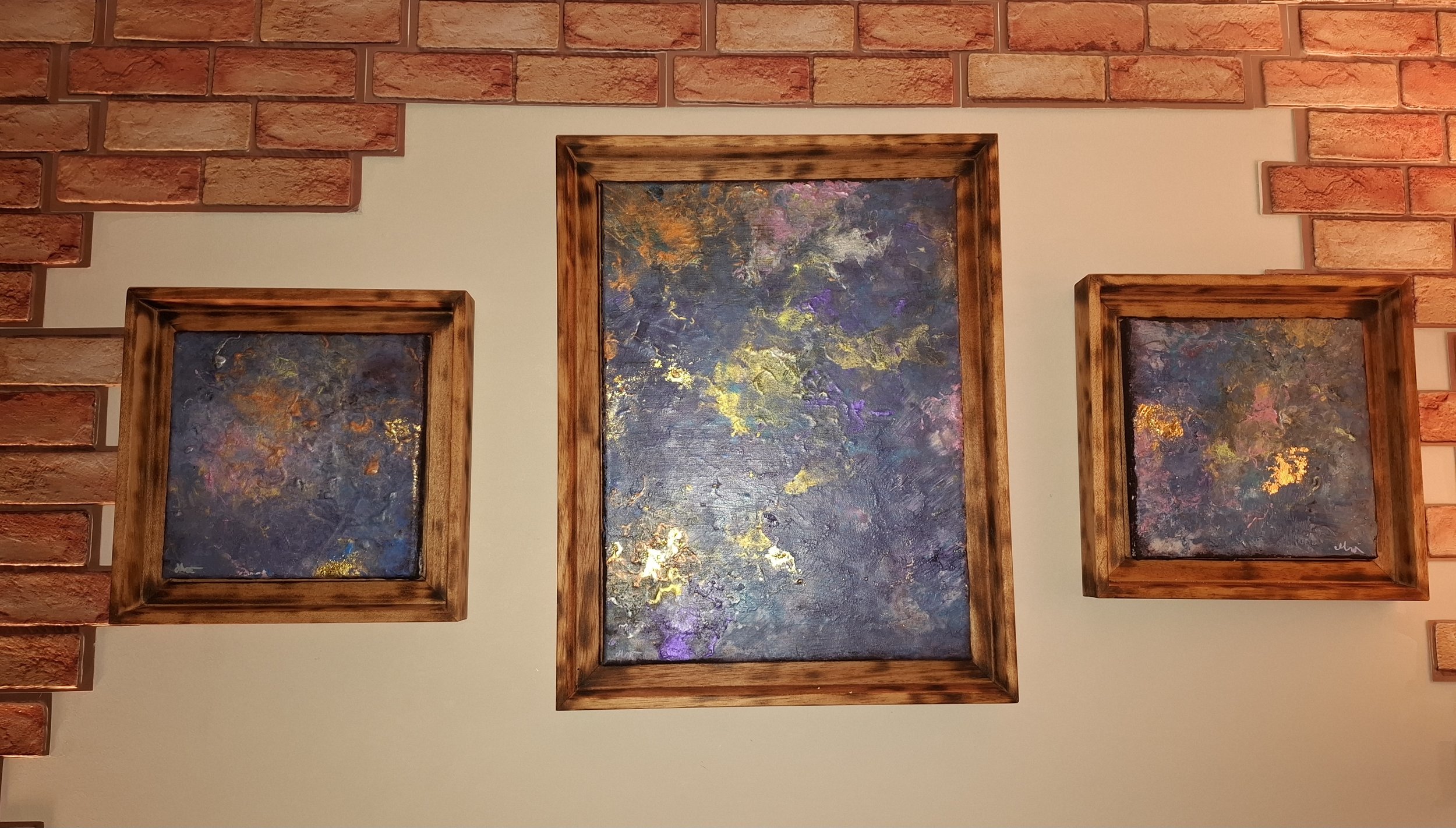 Drei abstrakte Gemälde mit bunten, goldfarbenen Akzenten in Holzbilderrahmen, an einer Wand mit Ziegelsteinmauer.