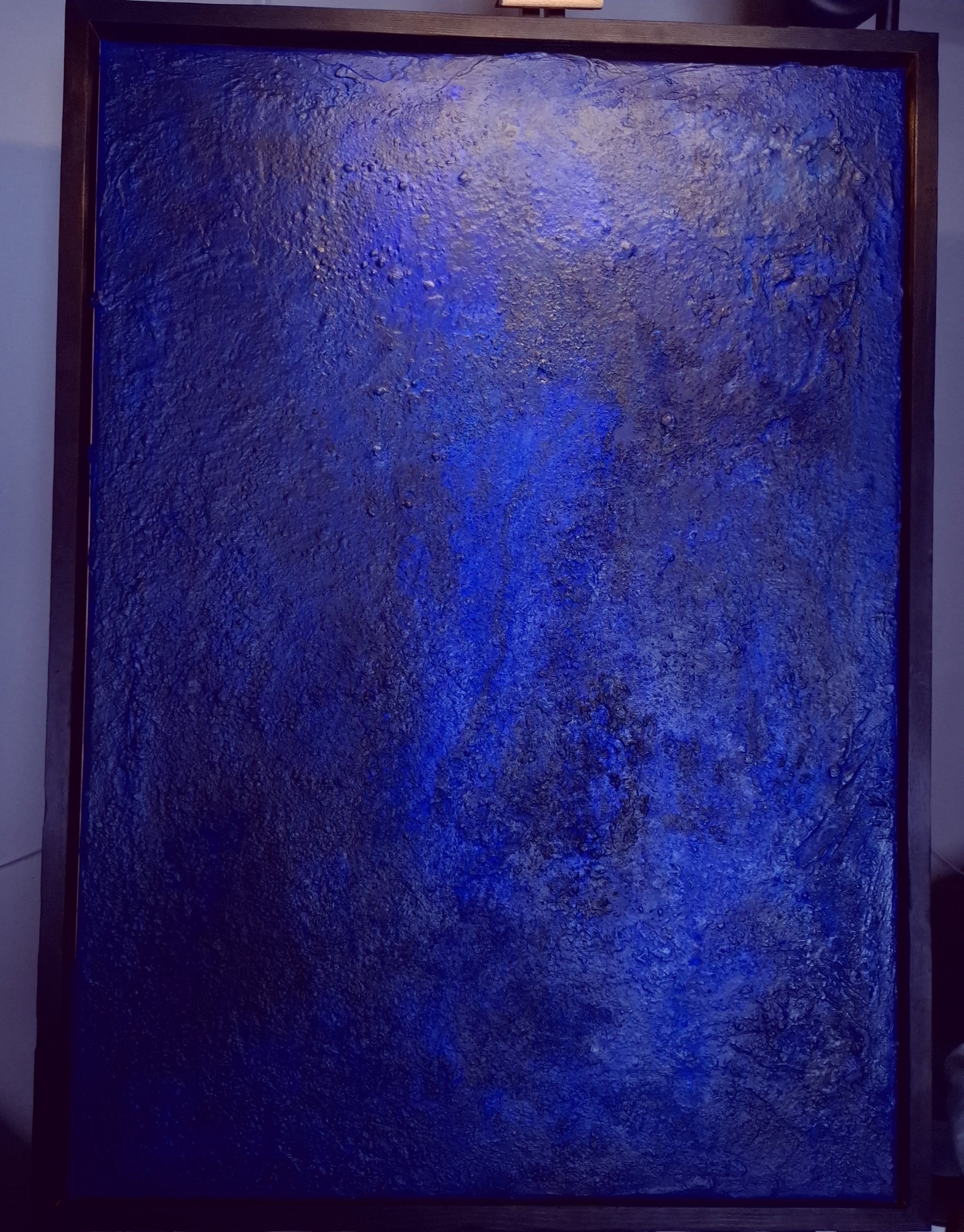 Ein großes, quadratisches Kunstwerk mit einer Oberflächenstruktur, die an Matsch oder Lava erinnert, wird dunkelblau beleuchtet und in einem Holzrahmen präsentiert.