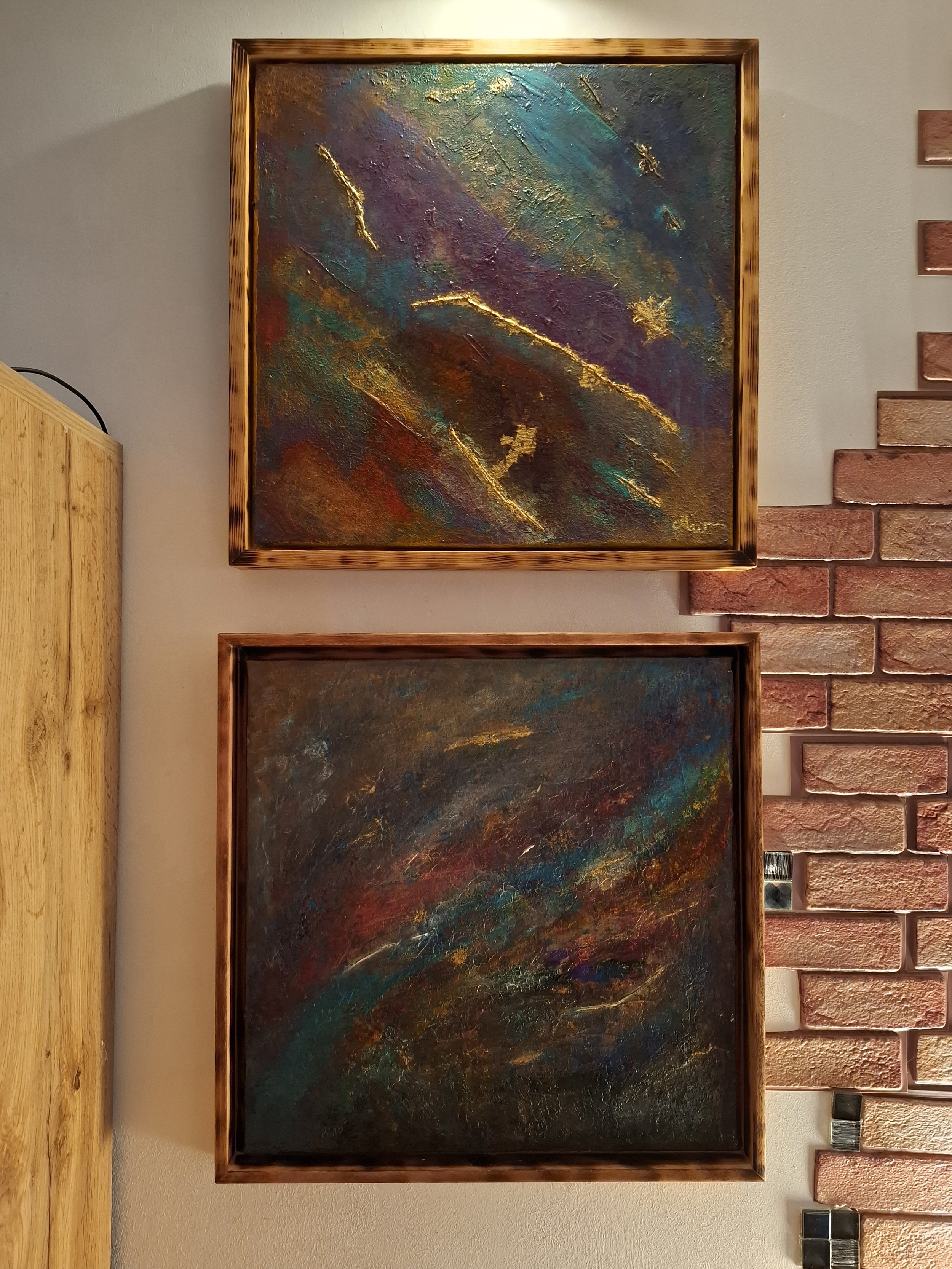 Zwei abstrakte Gemälde mit bunten, texturierten Flächen und goldenen Akzenten, hängen an einer weißen Wand neben einer roten Ziegelmauer.