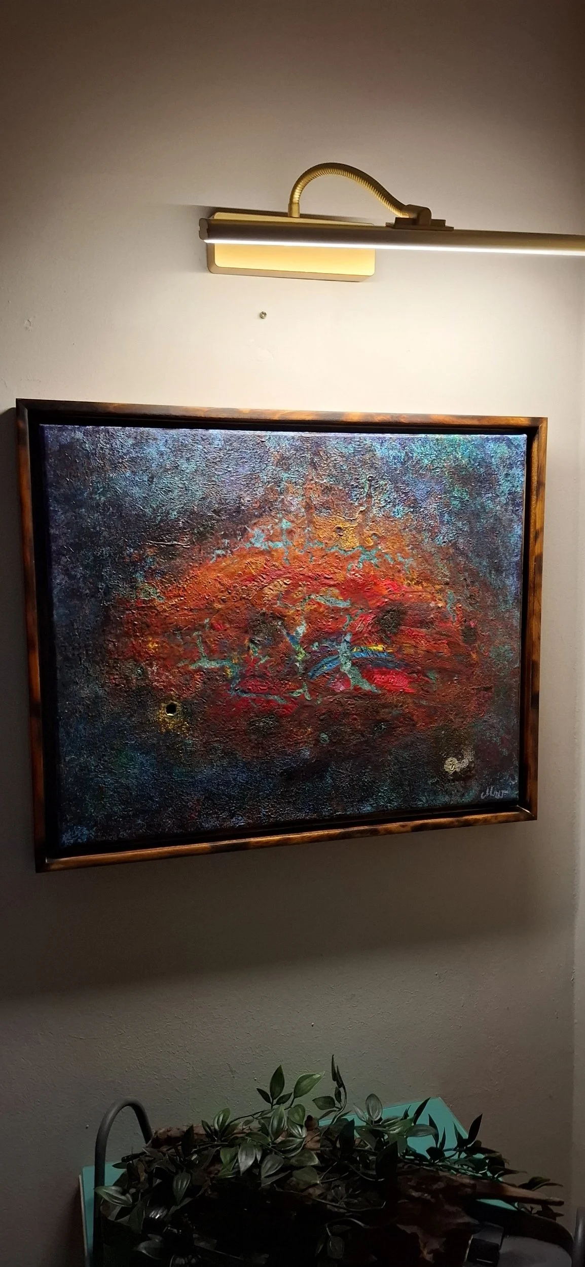 Ein modernes, abstraktes Gemälde mit rot, blau und orange Farbtönen, in einem dunklen Rahmen, an einer Wand beleuchtet mit einer goldenen Wandlampe.