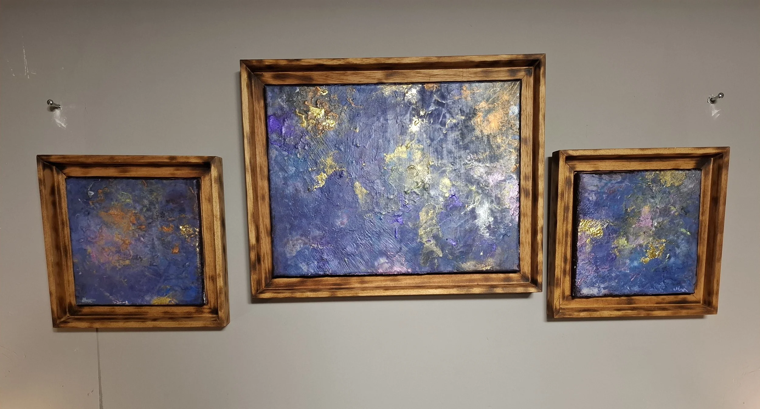 Drei abstrakte Gemälde mit blauen, lila und goldenen Akzenten, rahmenlos an einer weißen Wand aufgehängt.