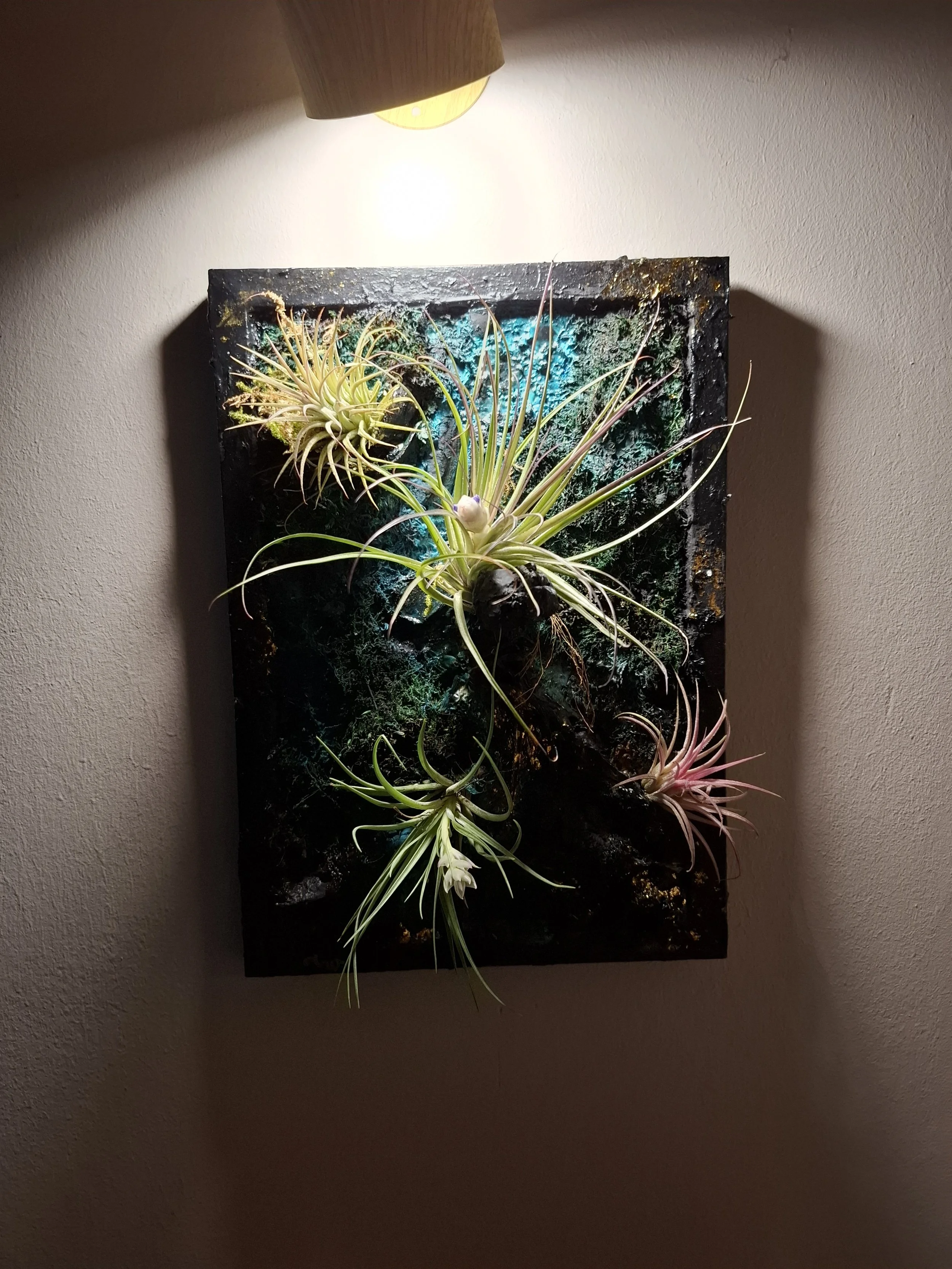 Kunstpflanzen in einem Wandbild unter einer Deckenlampe, mit dunklem Hintergrund und Lichtakzenten.