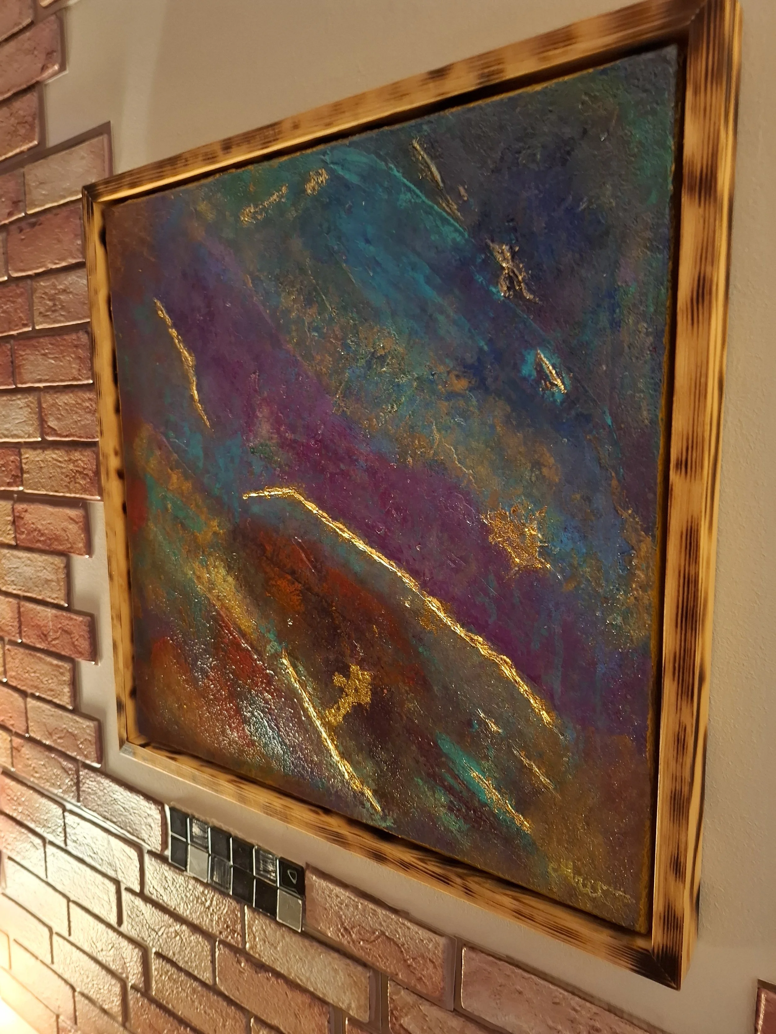 Abstraktes Gemälde mit bunten, goldbestickten Flächen innerhalb eines Holzbilderrahmens, an einer Ziegelsteinwand aufgehängt.