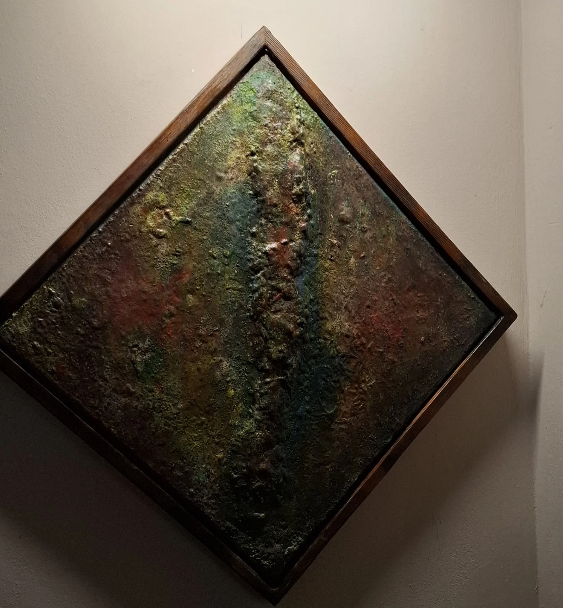 Ein abstraktes Gemälde mit tiefen Texturen und lebendigen Farben, gerahmt und an einer weißen Wand aufgehängt.