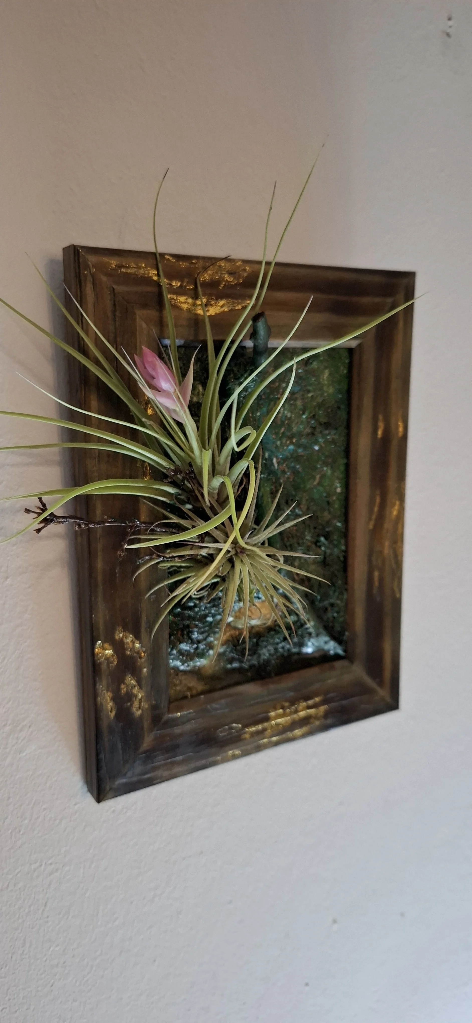 Ein Wandbild mit einem 3D- Pflanzenarrangement in einem Holzrahmen, das eine luftige Pflanze mit rosa Blume enthält.