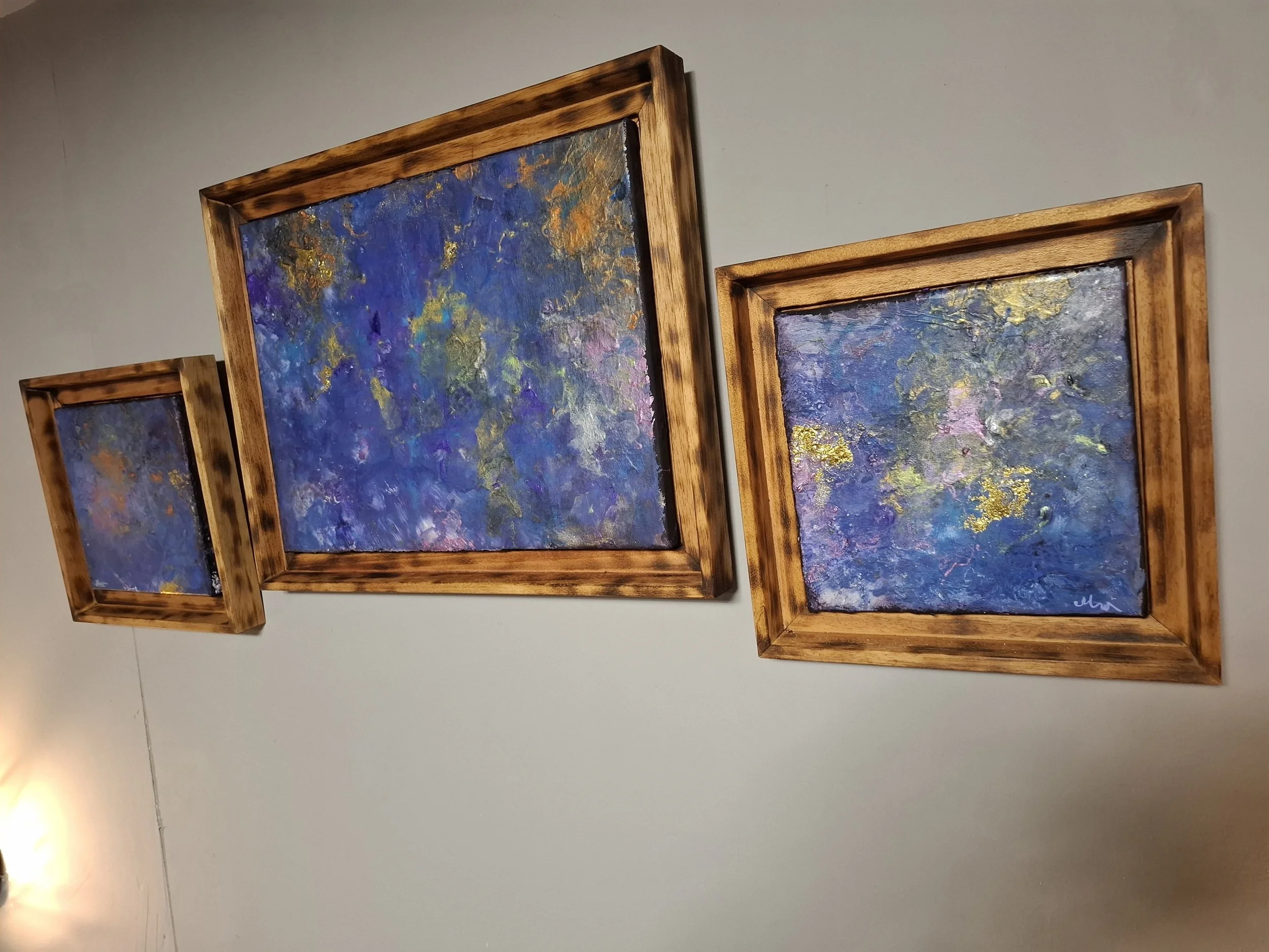 Drei abstrakte Gemälde in Holzrahmen, überwiegend in Blau- und Lilatönen, an einer weißen Wand