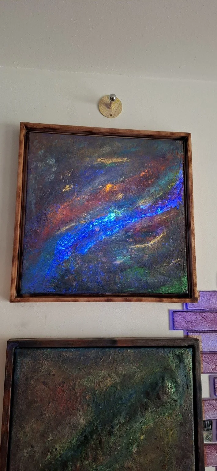 Zwei abstrakte Gemälde, eines mit dunklen, leuchtenden Farben und blauen Akzenten, das andere mit erdigen Tönen und grünen Elementen, hängen an einer Wand.