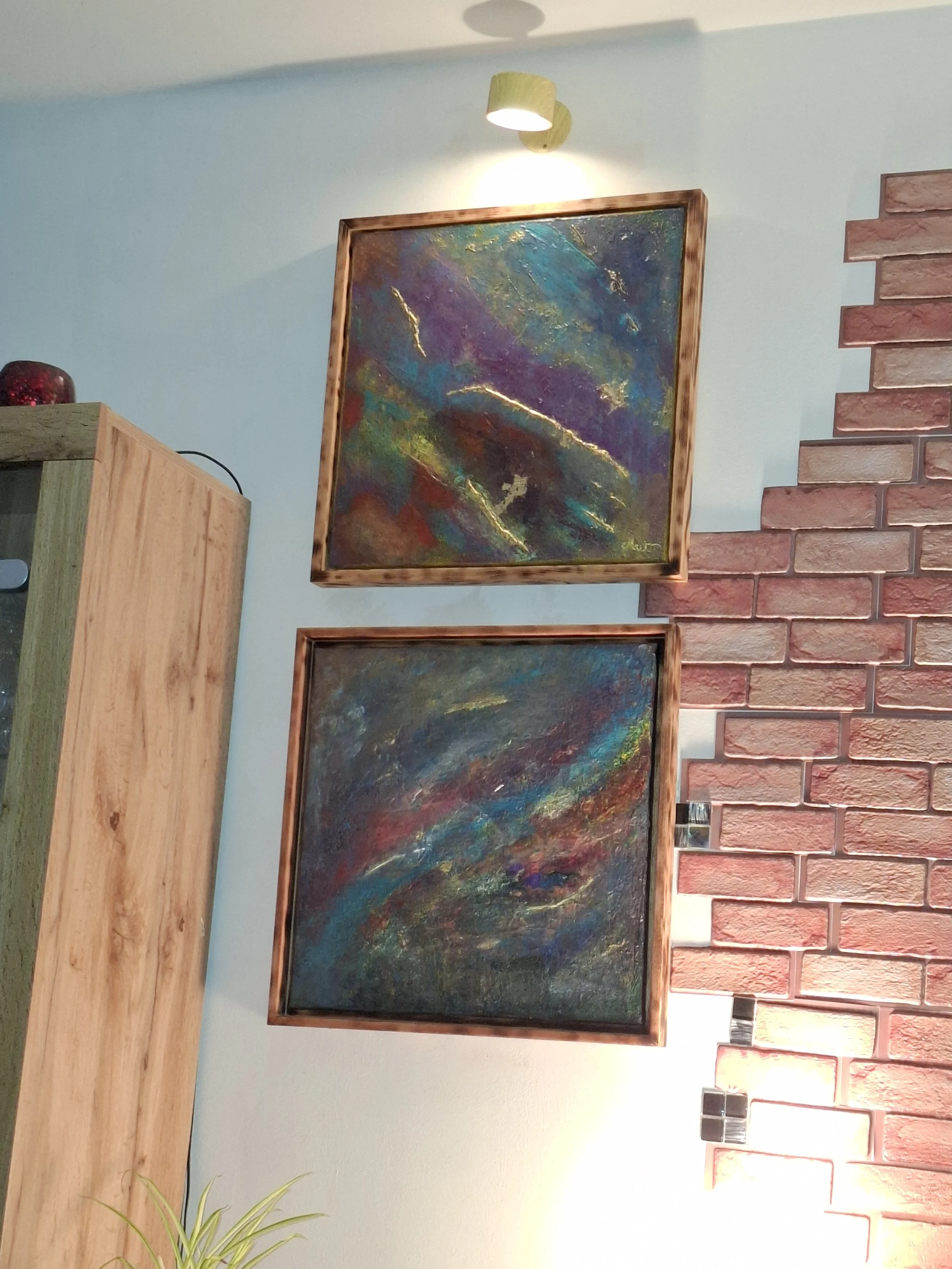 Abstraktes Gemälde mit dunklen, lebendigen Farben, goldenen Akzenten und einer Holzrahmung, an einer hellblauen Wand, beleuchtet von einer an der Wand angebrachten Stecklampe.