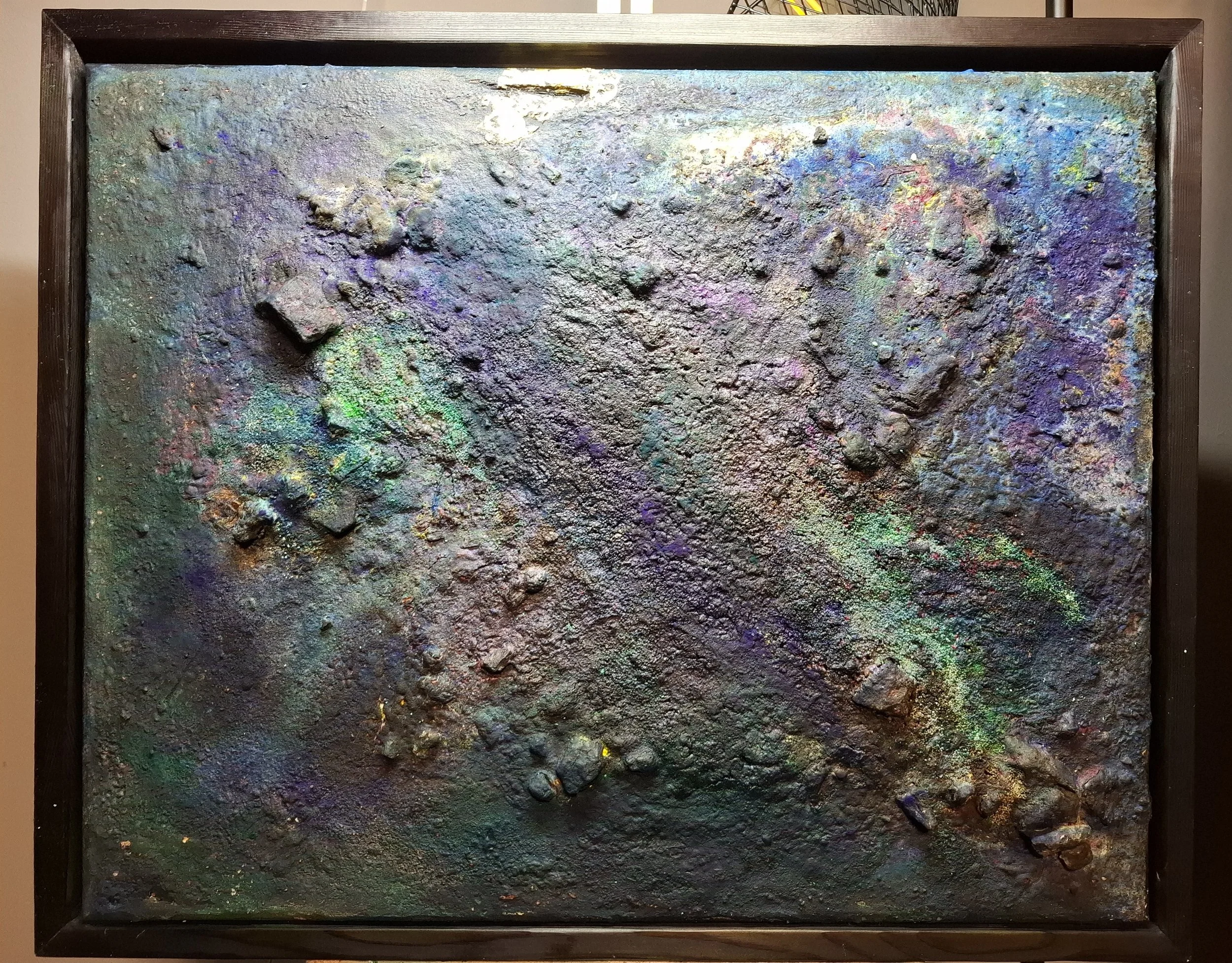 Eine Leinwand mit einer texturierten, metallisch schimmernden Oberfläche in dunklen Farbtönen mit irisiertem Farbreflex. Rahmen aus dunklem Holz umgibt das Kunstwerk.