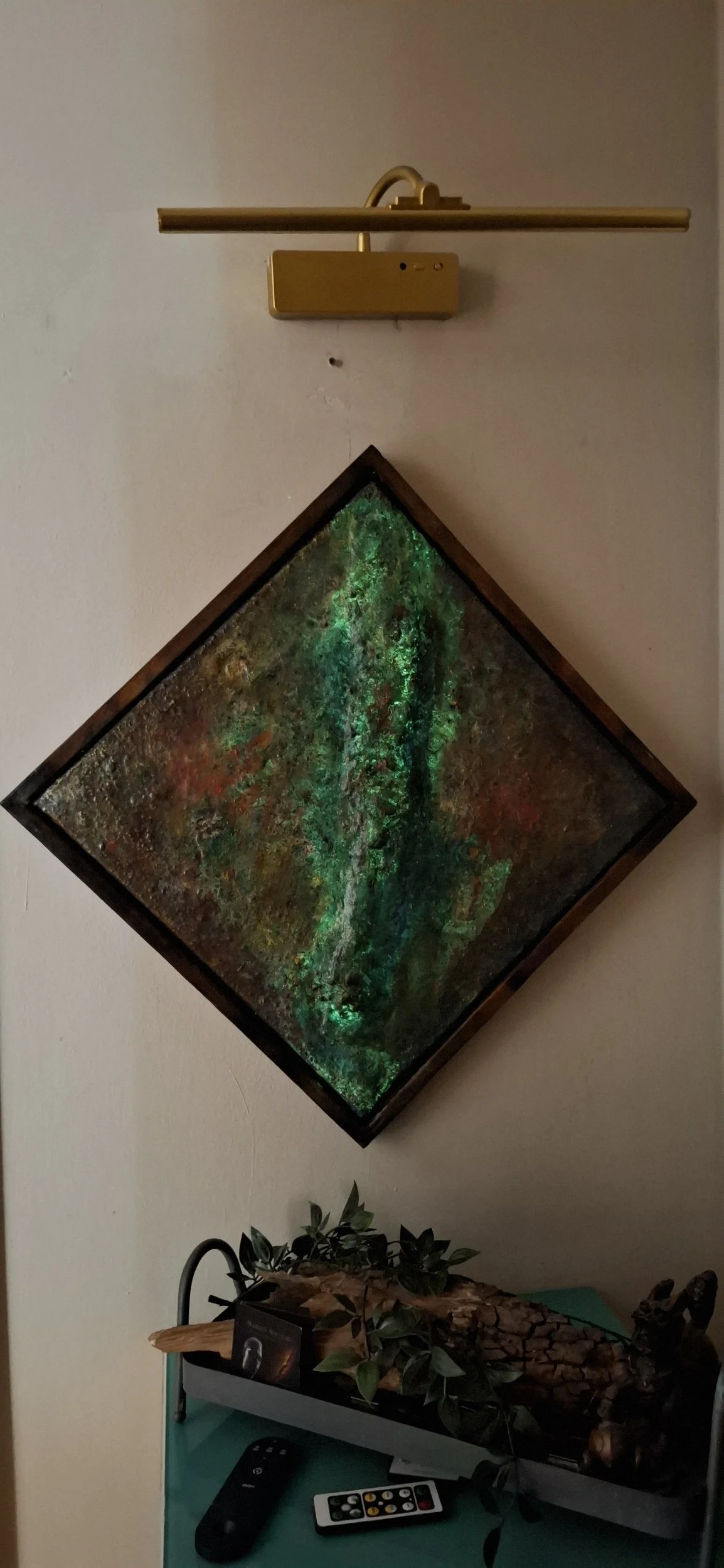 Abstraktes Gemälde mit grünen, roten und braunen Farben, diagonale Ausrichtung an der Wand, darunter eine kleine Tischfläche mit Pflanzen, Holzstücken und einem Fernbedienungs-Set.