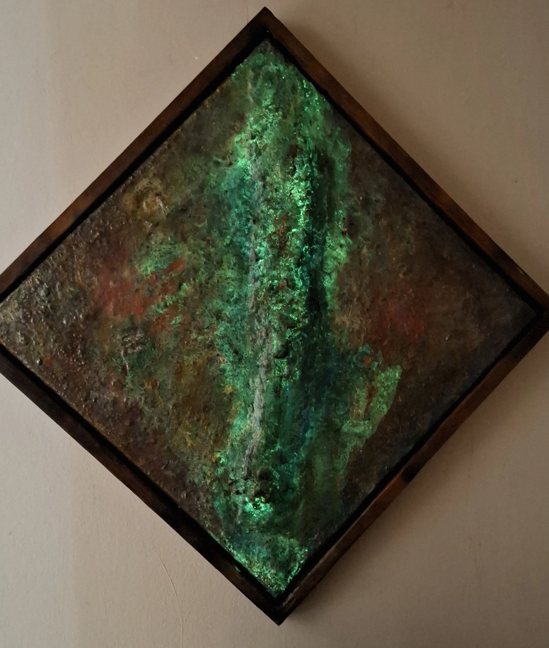 Ein quadratisches Kunstwerk im Rahmen, das aus einer Mischung aus bunten, schimmernden Materialien besteht, mit grünem Leuchteffekt, präsentiert an einer Wand.