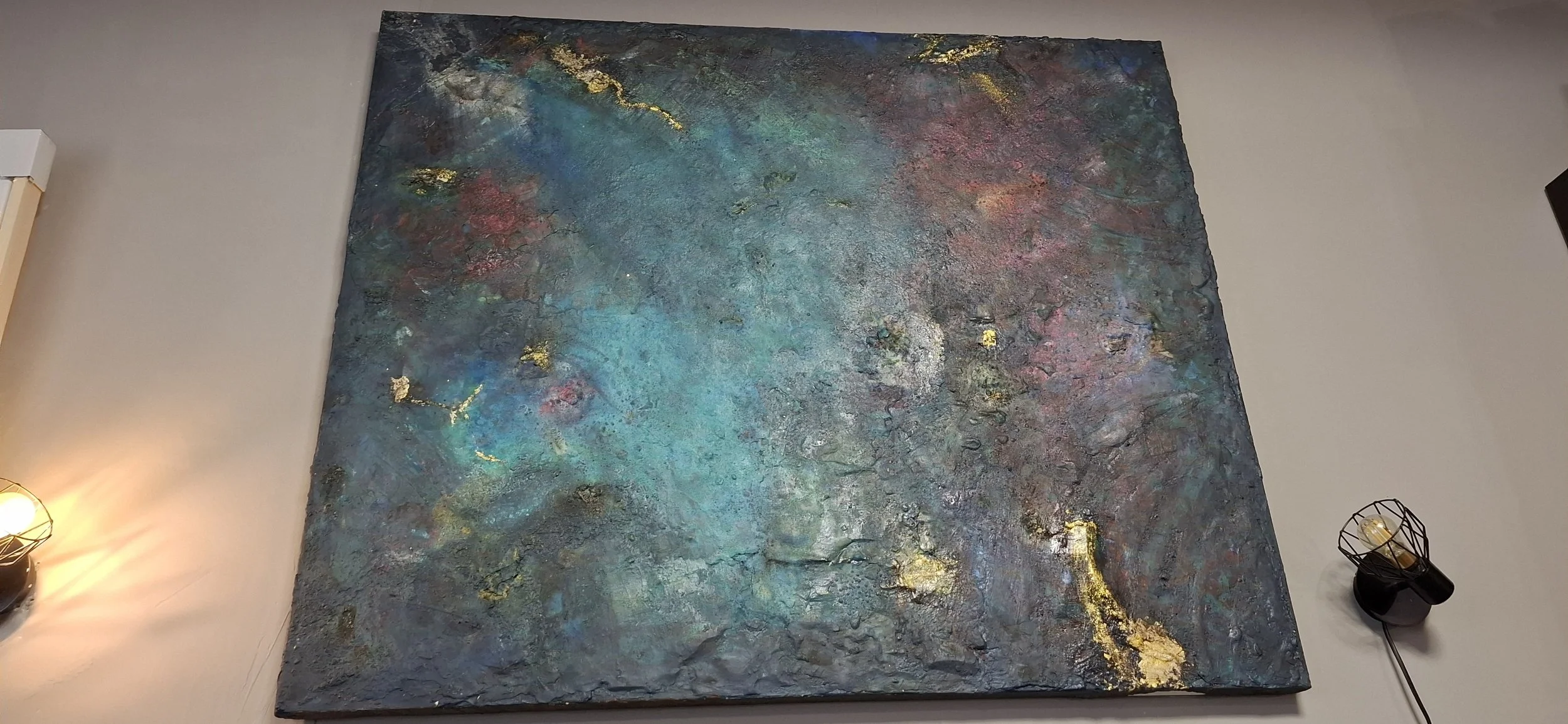 Ein abstraktes Gemälde mit blauen, grünen, roten und goldenen Akzenten, aufgehängt an einer weißen Wand, beleuchtet von seitlichen Lampen.