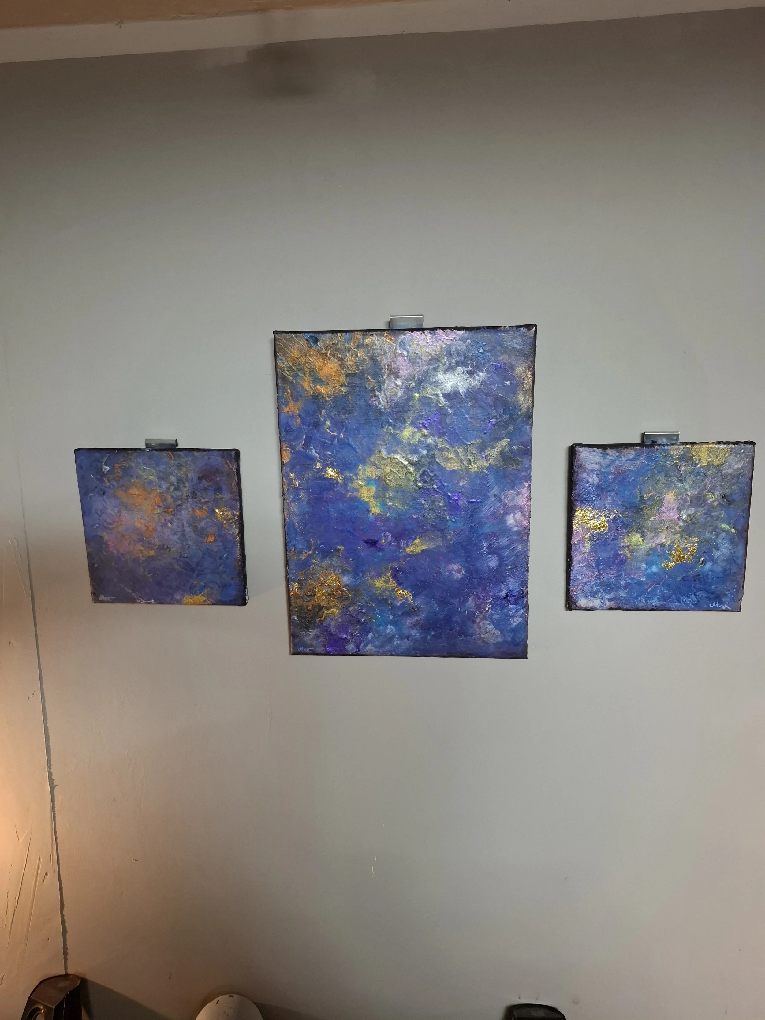 Drei abstrakte Gemälde in Blau, Lila, Gold und Weiß an einer weißen Wand. Das mittlere Gemälde ist größer als die beiden Seitengemälde.