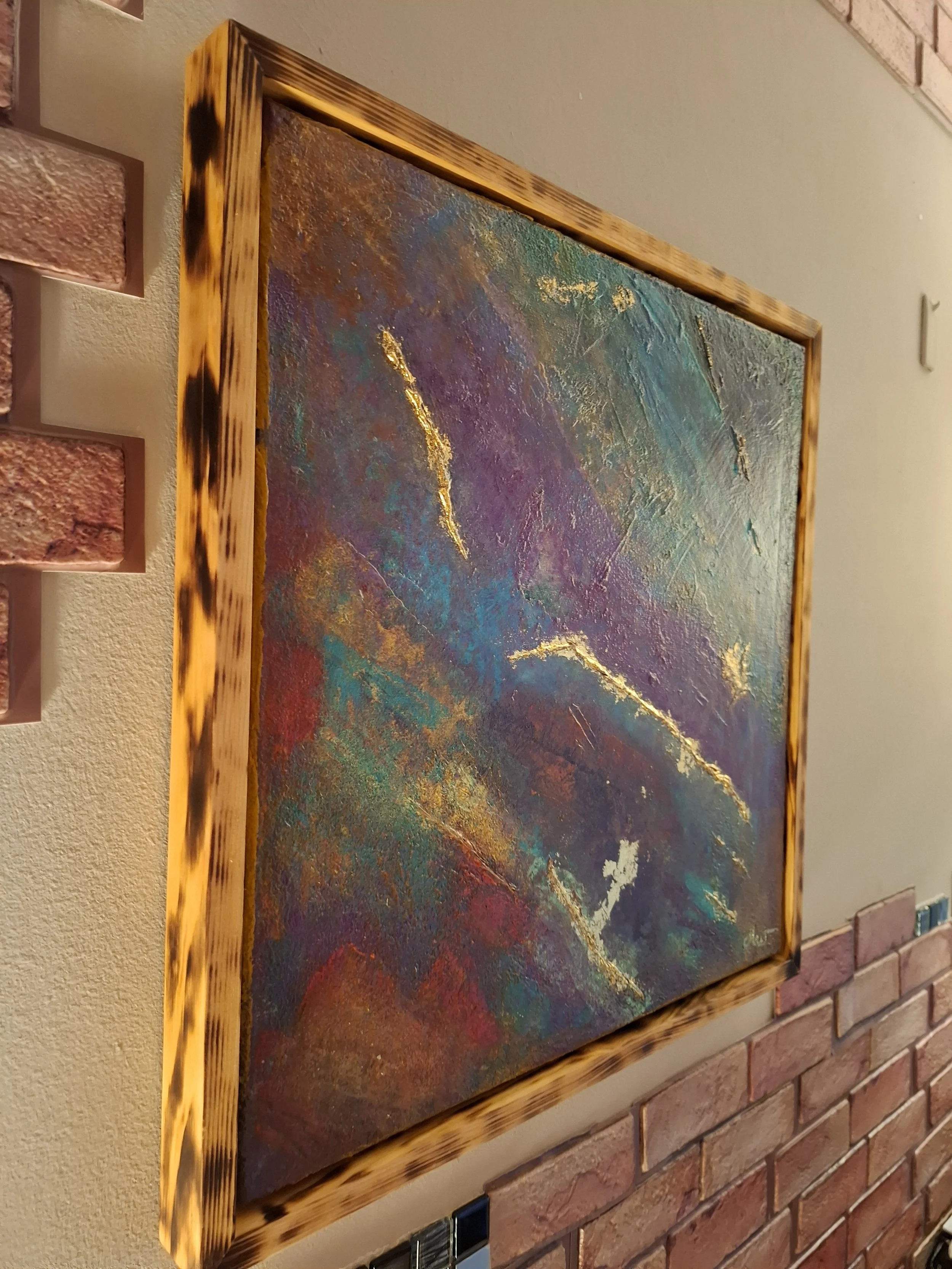 Abstraktes Gemälde mit goldenen Akzenten, eingerahmt in eine Holzrahmen mit Tigerfelsmuster, an einer Wand mit Ziegelstein und Putz.